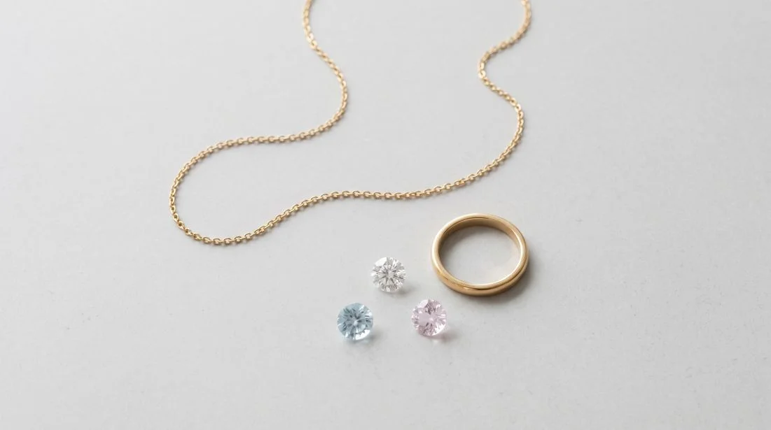 明るいグレーの石のテーブルに置かれた金色のチェーンネックレス、シンプルな金色の指輪、枠に留めていないカラーストーン数個